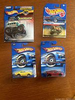 Set of 4 Hot Wheels cars in original sealed blister packaging on wooden surface. Includes one Monster Jam Surf Monster truck and three standard Hot Wheels cars.