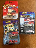 Three Johnny Lightning die-cast cars in blister packaging on a wooden surface, showing all three cars and their packaging designs.