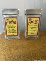 Front view of two vintage Planters peanut and almond glass canisters with metal lids and yellow labels.