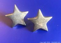 Front view of two silver star-shaped earrings against a blue background, showing star shape and surface texture.