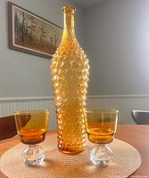 Amber Italian glass genie bottle with pineapple texture pattern, placed between two amber glass goblets with clear bubble bases on a round woven placemat on a wooden table.