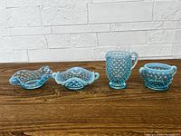 Four piece set of vintage Fenton blue opalescent hobnail glass cream and sugar items placed on wooden surface with white textured brick background.