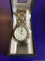 Front view of the watch in its box showing white dial with gold Roman numerals and gold-accented bezel and band.