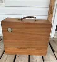 Closed walnut sewing machine case, front-facing with Singer emblem and leather handle on top.