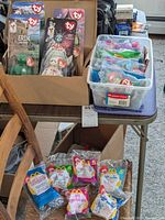 Photo shows four TY Beanie Babies in packaging standing in a box behind a plastic container filled with multiple sealed Happy Meal toys in plastic bags on a table.