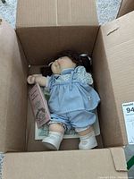 Front and top views of the Cabbage Patch doll inside an open cardboard box, showing facial details, clothing, and certificates.
