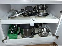 Full view of cookware items arranged in a cabinet shelf, showing various pots, pans, strainers, and lids.