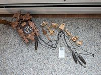 Photo showing German Black Forest style wooden cuckoo clock with carved leaf and bird decorations, laying on carpet, along with attached pine cone weights and multiple small wooden miniature doll figures with yarn hair scattered around it.