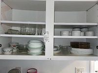 Wide view of kitchen cabinet showing stacks of Corelle plates with green trim, floral patterned bowls, clear glass bowls, and white ramekins neatly arranged on two shelves.