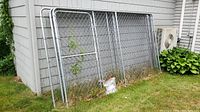 Unassembled chain link dog kennel panels leaning against outdoor wall, showing weathering and minor grass debris at bottom.