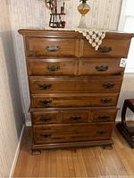 Front view of the tall wooden dresser with six drawers and metal handles, showing the full piece against a wall with a doily and decorative items on top.