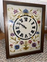 Front view of rectangular wooden framed clock with hand embroidered fabric face showing Roman numeral dial and floral embroidery.