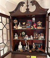 Cabinet open showing three glass shelves filled with various figurines, music boxes, and collectibles including carousel music box, nutcracker figure, cat figurine, and thimbles.