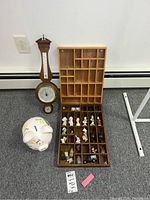 Wide shot of the lot showing two wood shadow boxes filled with figurines, a wall-mounted barometer, and a white painted ceramic piggy bank on carpeted floor.