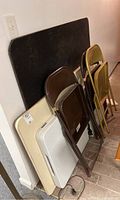Three folding tables stacked against a wall with three folded metal chairs placed alongside them. Tables are beige and white with visible scratches.