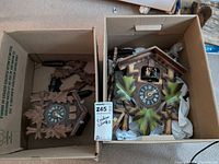 Two cuckoo clocks side by side in cardboard boxes: unpainted carved wood clock on left, painted chalet style clock on right