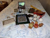 Full overview of the lot items spread out on a bed showing the variety of collectibles including figurines, framed stamps, beaded bag, jewelry box with assorted jewelry, ceramic balls, and collector tin.