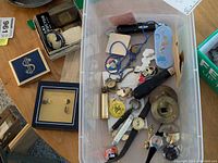 Photo showing a plastic bin with assorted wristwatches, pins, a carved cameo brooch, locket pendant, finger cymbals, and a small metal box. Sterling silver earrings displayed in separate box on table.