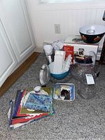 Full view of lot on floor showing food dehydrator box, glassware, plastic bowls, water filter pitchers, and kitchen tools.