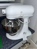 White KitchenAid Ultra Power stand mixer with a stainless steel mixing bowl attached and three attachments visible inside the bowl: wire whisk, dough hook, and flat beater. Mixer has a control knob with speed settings from 0 to 10.