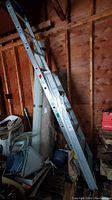 Photo showing the taller aluminum extension ladder leaning against the wall in a garage with surrounding items.