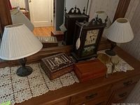 Wide view showing vintage mantle clock, two metal base lamps with white pleated shades, wood inlay box, Lane cedar wood box, and vintage hand mirror and brush on lace runner.