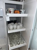 View of three pull-out shelves in a white cabinet containing mugs on the top two shelves and various clear drinking glasses on the bottom shelf.