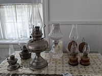 Seven oil lamps displayed on a lace-covered table including a pair of Nasco Handy Camp oil lamps, Rayo oil lamp, Lamp Light oil lamp, and others with various glass chimneys and metal bases.