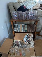 Photo showing 18 New England Patriots logo glasses on wooden side table with Boston Red Sox baseball cap on top, and a cardboard box containing additional glasses below.
