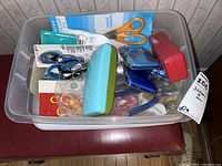 Photo showing bin with orange handled scissors, blue handled scissors, rotary cutter, stapler, metal case containers, sewing hooks, and fasteners.