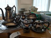Photo showing silver plate coffee pot, teapot, cream and sugar bowls, serving bowls, and part of porcelain plates and cups stacked in background.