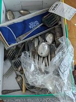 Photo showing an assortment of silver plated forks, spoons, a slotted spatula, and other utensils in a box, some partially wrapped in plastic.
