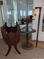 Display of round 2-tier wooden table with carved duck head on upper tier, standing next to carved wood walking cane and wooden magazine rack with scalloped edges