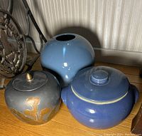 Three ceramic pottery pieces on wooden surface, including one light blue round vase, one dark blue lidded jar, and a gray vase with gold floral design and lid.
