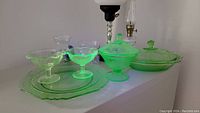Uranium glass covered dishes, dessert bowls, plates, glass vases with beads, and two oil lamps shown under normal light, displayed on white surface.