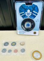 Photo showing a sealed set of Canadian coins alongside a Toronto Maple Leafs mini hockey jersey and puck in package, plus the silver Flamingo Hilton $10 token separate on table.