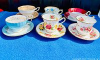 Six bone china vintage cup and saucer sets displayed on blue fabric base, showing diverse floral patterns and gold trim.