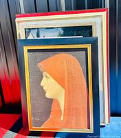 Framed color portrait of woman in orange hooded garment in profile with black and gold frame.