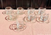 Seven clear glass cups and saucers arranged in two groups on a surface, showing two styles of vintage glassware.
