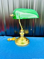 Front angle view of the bankers desk lamp showing the green opaline glass shade and brass base on a blue cloth surface with a dark background.