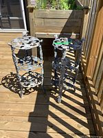 Two aluminum plant stands with ornate floral designs and three tiers, placed on a wooden deck with sunlight and shadows.