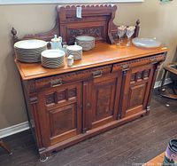 Front view of antique wooden sideboard with stacked dishes, cups, and glasses on top. Visible carved wood details and drawer/cupboard handles with keys.