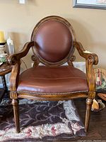 Front view of arm chair showing brown leather-like padded round backrest and seat with brass nailhead trim, wooden carved arms, and legs.