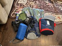 Photo showing all items in the lot arranged on a hardwood floor and a carpet nearby, including blue rolled blanket, bags, cases, and binoculars.