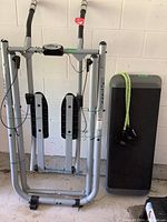 Photo of a silver-grey Gazelle rowing machine with black foot pedals and handlebars standing next to a black exercise platform and a green stretching rope within a garage setting.
