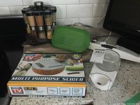 Photo shows multi purpose slicer box, spice rack with six jars, green storage container with lid, and white mandolin slicer on kitchen counter.