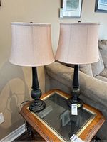 Two matching lamps with dark metal bases and beige fabric shades standing on a glass-top wooden table beside a sofa.