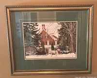 Framed print showing winter scene with horse-drawn sleigh and brick house framed in gold-tone frame with green mat.