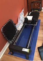 Full view of reformer frame on blue mat showing carriage, foot bar, rails and rebounder accessory
