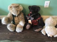 Three plush bears seated on dark wood floor against light green wall. Bears vary in size and color with distinct clothing accessories.